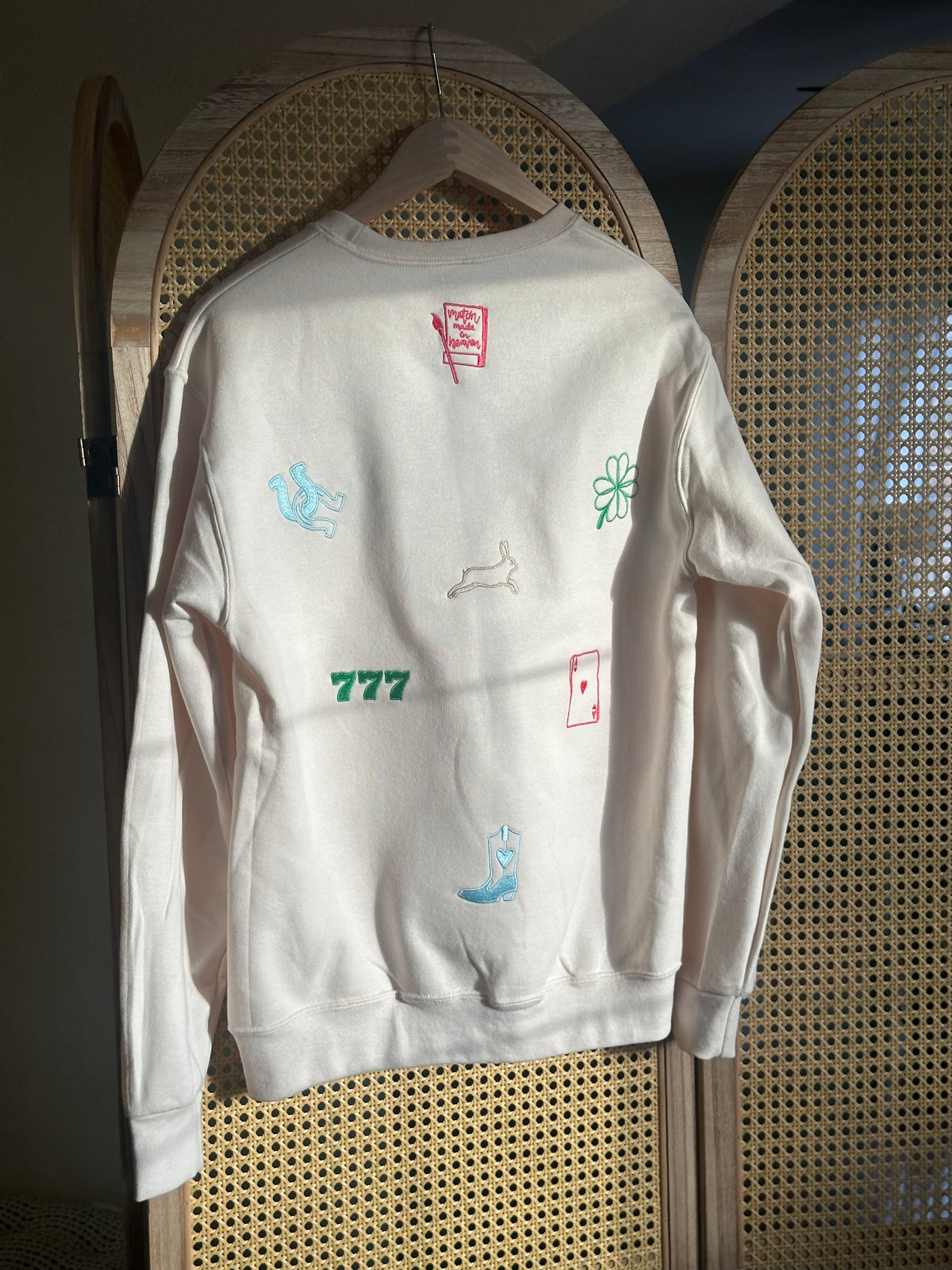 Back view of a cream crewneck featuring colorful embroidered St. Patrick’s Day-inspired lucky symbols, including horseshoes, a rabbit’s foot, playing cards, cherries, and “777” lettering.