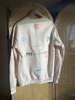 Back view of a cream crewneck featuring colorful embroidered St. Patrick’s Day-inspired lucky symbols, including horseshoes, a rabbit’s foot, playing cards, cherries, and “777” lettering.