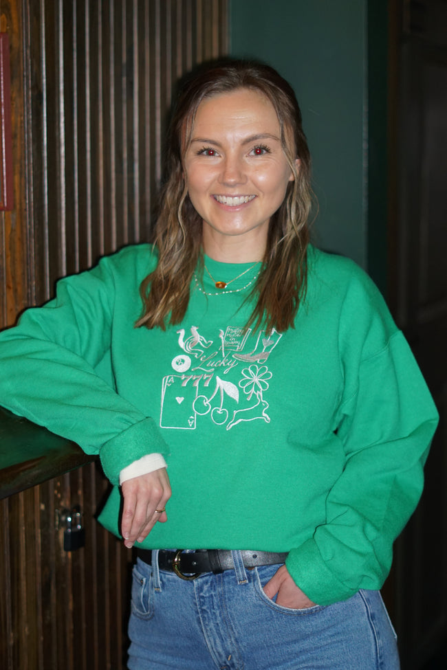 Woman wearing a green crewneck sweatshirt with embroidered Lucky Girl icon embroidery.