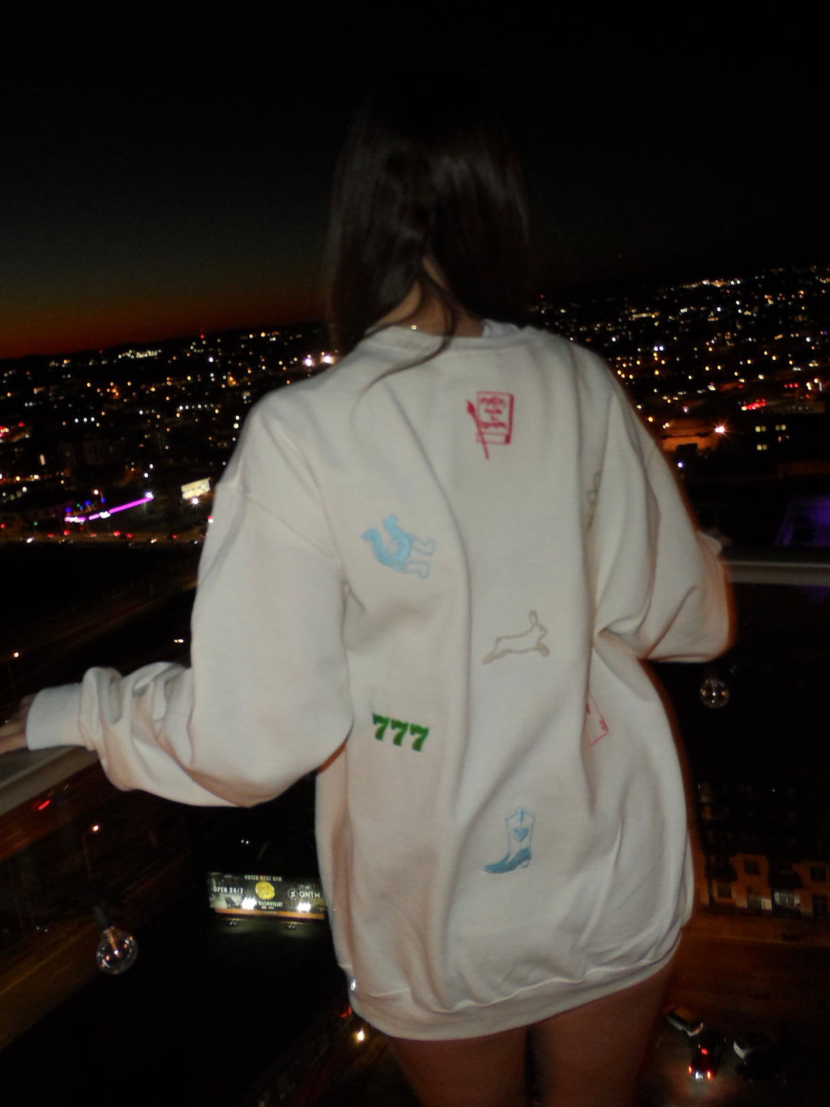 Back of a woman wearing a white embroidered hoodie with horseshoes, lucky numbers, cherries, cards, and other good luck-inspired designs, paired with denim shorts.