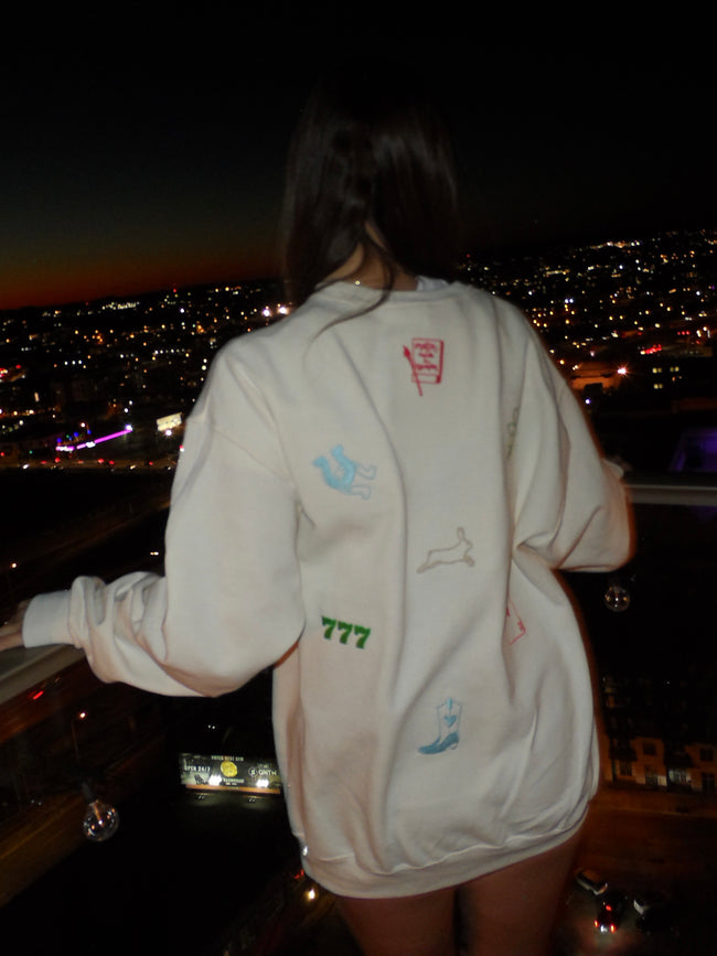 Back of a woman wearing a white embroidered hoodie with horseshoes, lucky numbers, cherries, cards, and other good luck-inspired designs, paired with denim shorts.