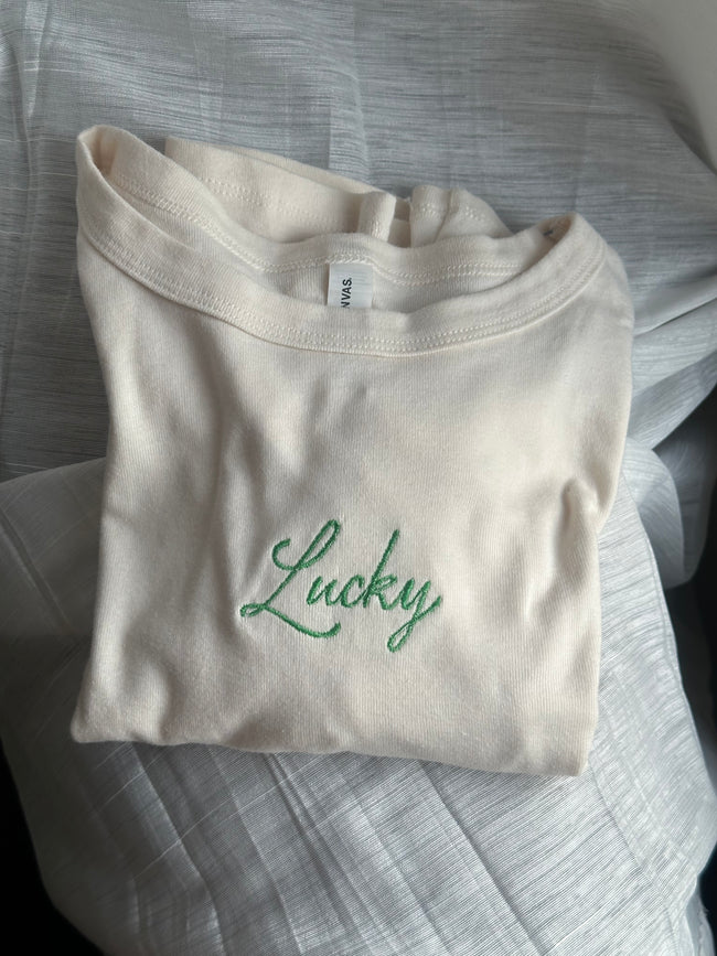 Ivory baby tee featuring green embroidered “Lucky” lettering across the chest.