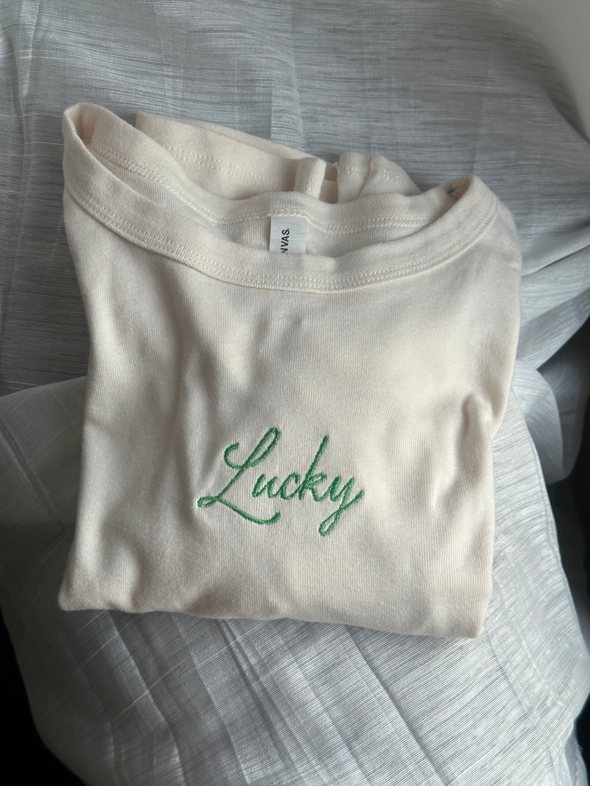 Ivory baby tee featuring green embroidered “Lucky” lettering across the chest.