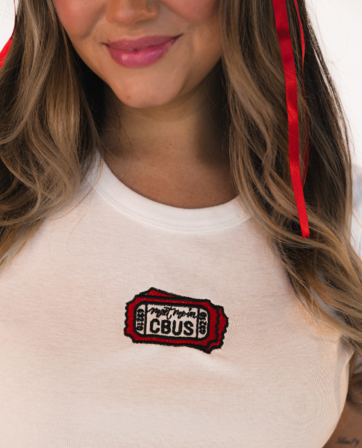 Woman wearing a white fitted baby tee featuring embroidered “Meet Me in CBUS” ticket design on the chest.
