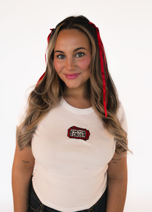 Woman wearing a white fitted baby tee featuring embroidered “Meet Me in CBUS” ticket design on the chest.