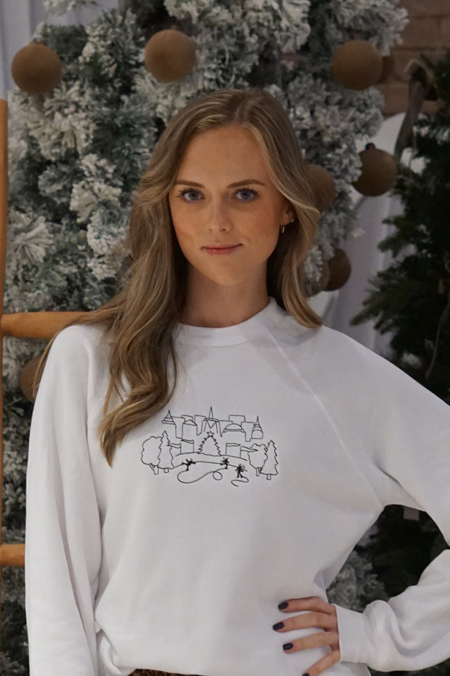 Woman wearing a white embroidered NYC Holiday crewneck sweatshirt featuring a winter city design.