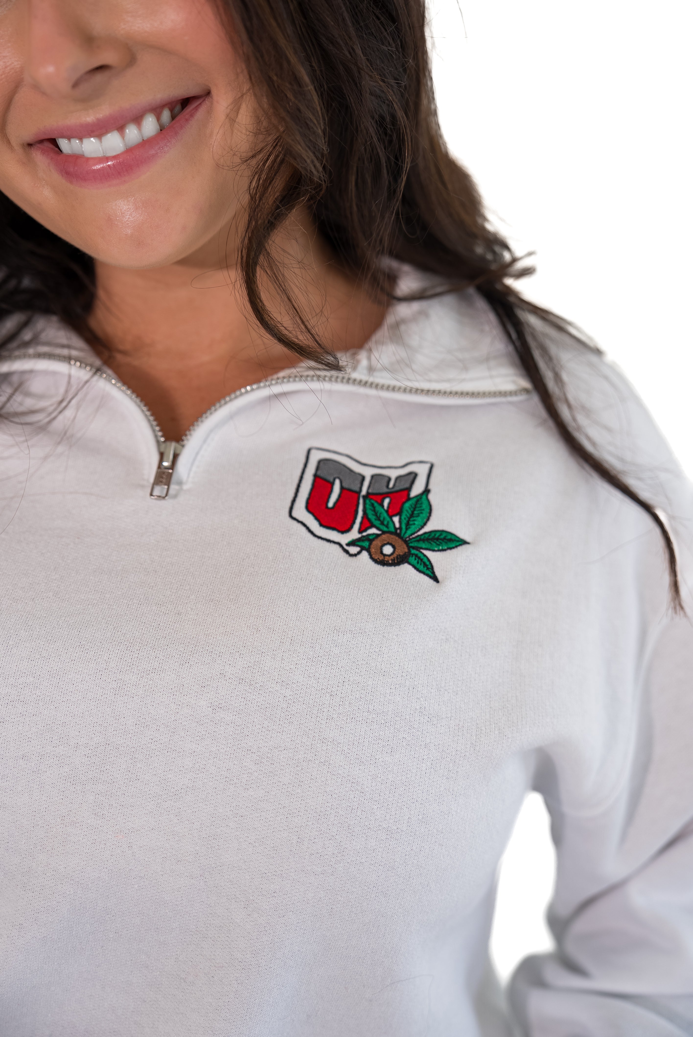 A close-up of a woman wearing a white half-zip pullover sweatshirt with a red, white, and green Ohio logo embroidered on the left chest.