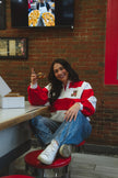 A woman sitting on a red stool in a cafe, wearing a red and white striped quarter-zip sweatshirt with a teddy bear patch on the chest, blue jeans, and white sneakers.