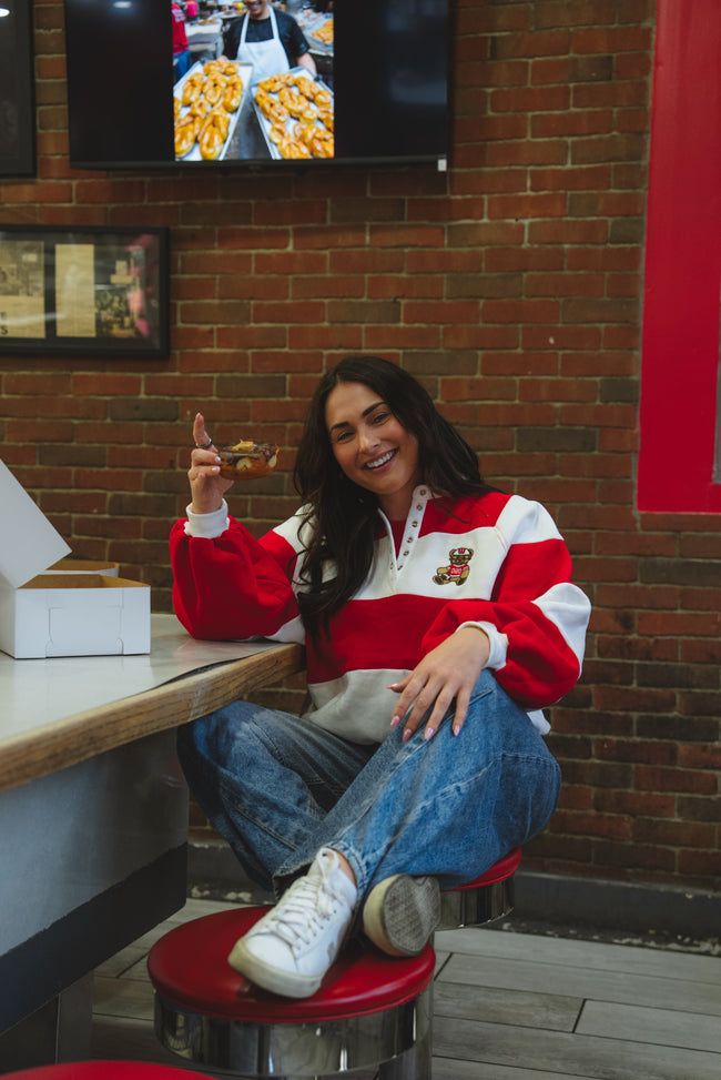 A woman sitting on a red stool in a cafe, wearing a red and white striped quarter-zip sweatshirt with a teddy bear patch on the chest, blue jeans, and white sneakers.