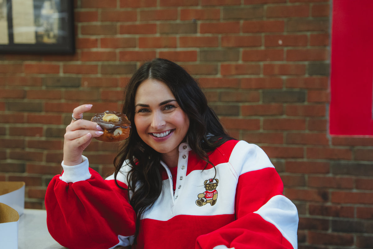 A woman smiling and holding a donut in front of her face, wearing a red and white pullover sweatshirt with a teddy bear graphic on the chest, set against a brick wall background.