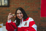 A woman smiling and holding a donut in front of her face, wearing a red and white pullover sweatshirt with a teddy bear graphic on the chest, set against a brick wall background.