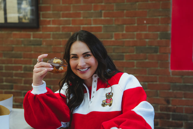 A woman smiling and holding a donut in front of her face, wearing a red and white pullover sweatshirt with a teddy bear graphic on the chest, set against a brick wall background.
