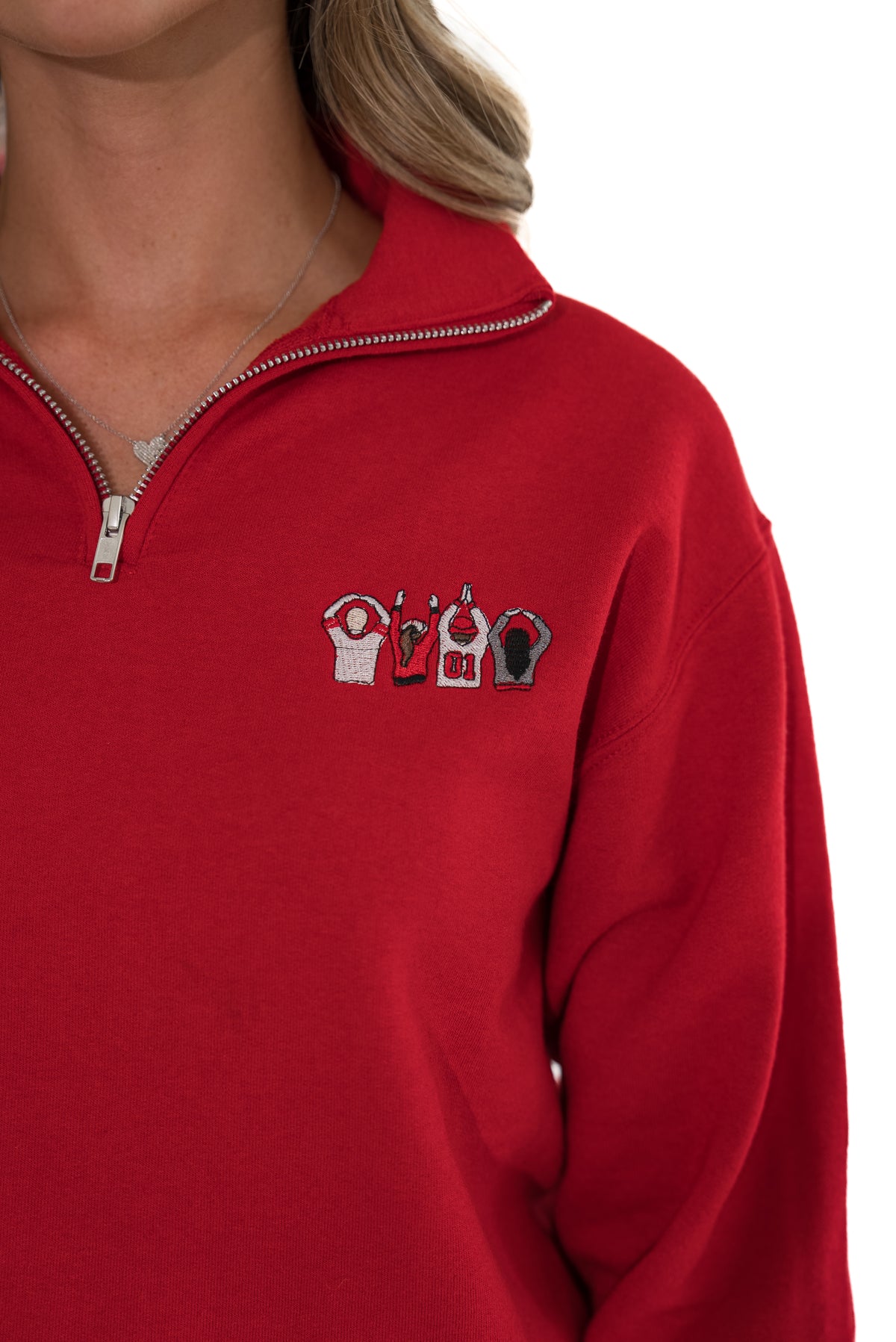 A close-up of a person wearing a bright red quarter-zip sweatshirt. The chest features a small embroidered graphic of four stylized figures in white, red, and black.