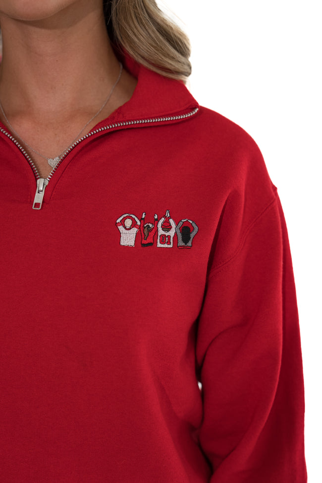 A close-up of a person wearing a bright red quarter-zip sweatshirt. The chest features a small embroidered graphic of four stylized figures in white, red, and black.