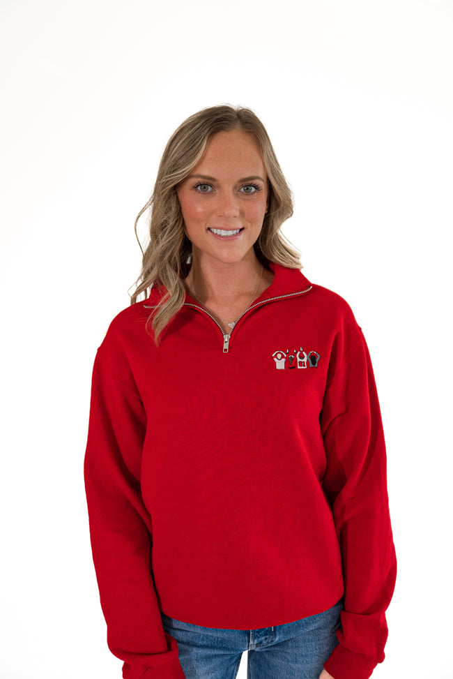 A smiling blonde woman wearing a bright red quarter-zip sweatshirt with a small embroidered logo on the left chest, paired with blue jeans, against a plain white background.