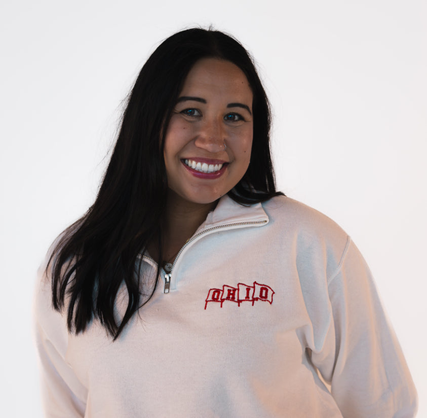 Woman wearing a white quarter zip sweatshirt with red embroidered flags, spelling 
