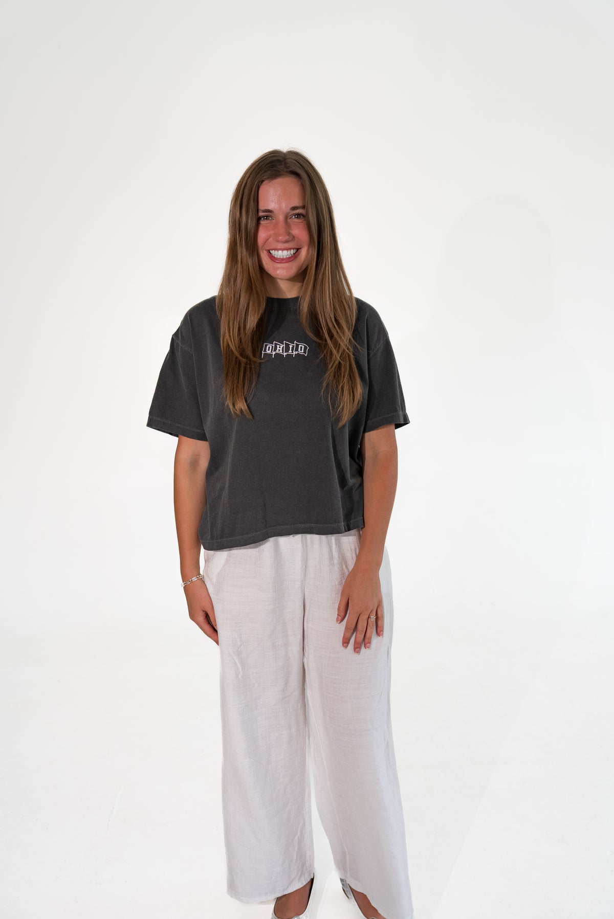 A woman smiling while wearing a dark charcoal gray graphic t-shirt and light beige wide-leg linen pants against a white studio background.