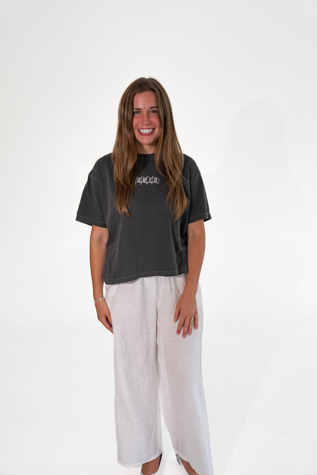 A woman smiling while wearing a dark charcoal gray graphic t-shirt and light beige wide-leg linen pants against a white studio background.