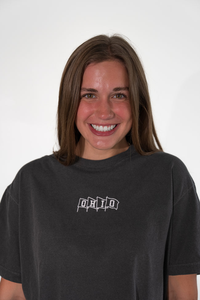 A smiling woman with shoulder-length brown hair wearing a faded black t-shirt with a white 'OHIO' graphic on the chest, set against a plain light gray background.