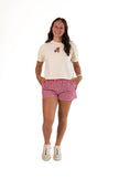 Smiling woman standing against a white background, wearing a cream-colored short-sleeve graphic tee with an embroidered Ohio football bear patch, red and white gingham lounge shorts, and sneakers.