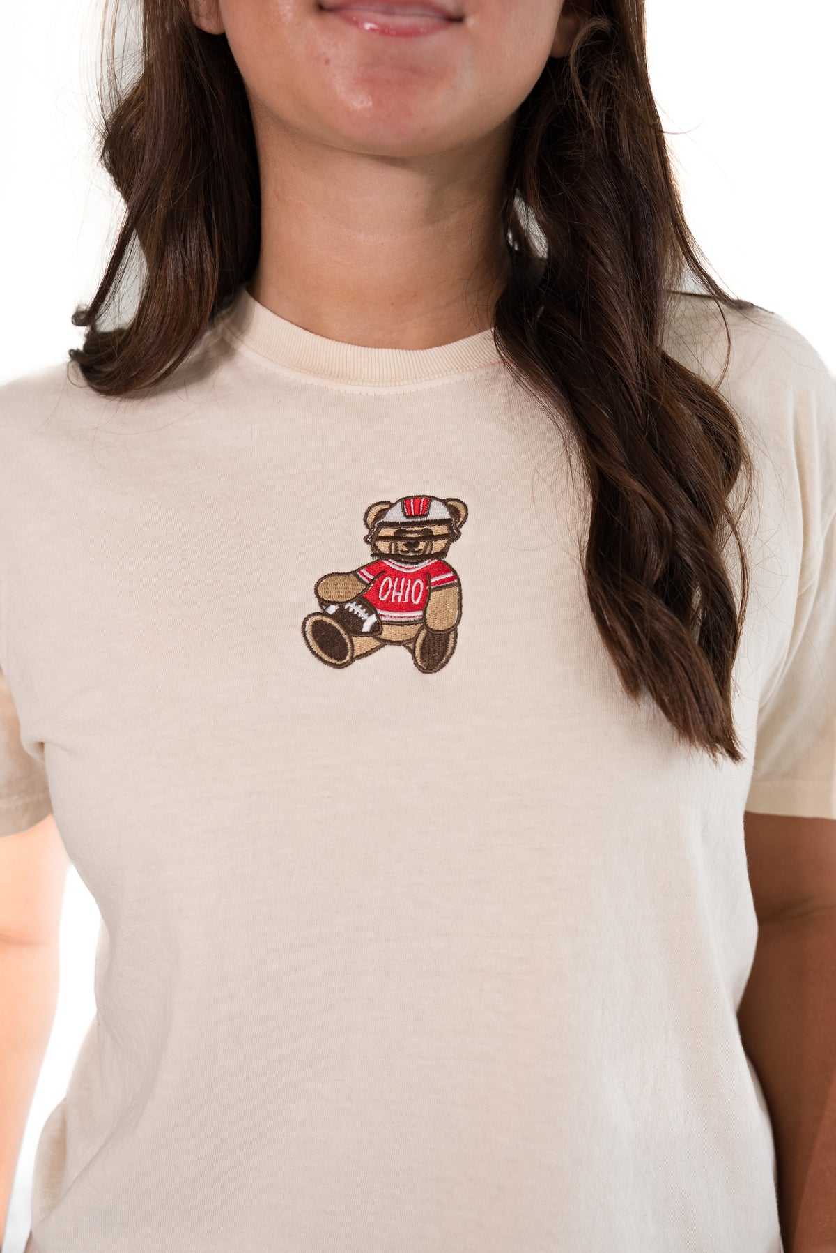 A close-up of a woman wearing a cream-colored t-shirt with a small embroidered teddy bear graphic on the chest. The teddy bear is brown with a red shirt that says 'OHIO'.