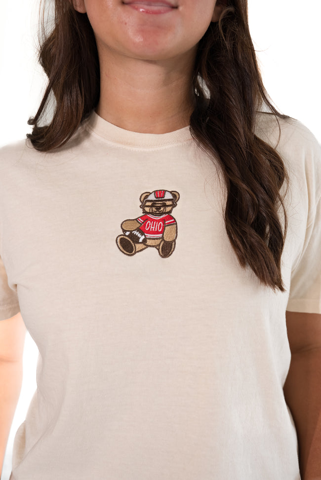 A close-up of a woman wearing a cream-colored t-shirt with a small embroidered teddy bear graphic on the chest. The teddy bear is brown with a red shirt that says 'OHIO'.