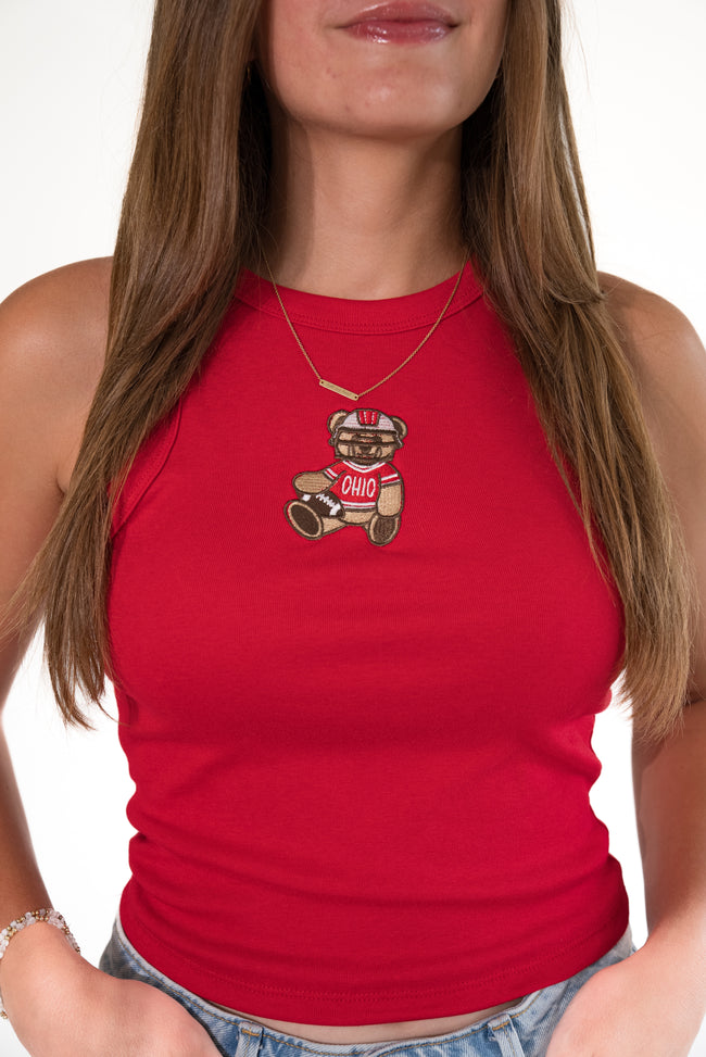 Woman wearing a bright red ribbed high-neck tank top featuring a stitched teddy bear patch on the chest, dressed in an Ohio football uniform and holding a football, paired with light-wash denim jeans.