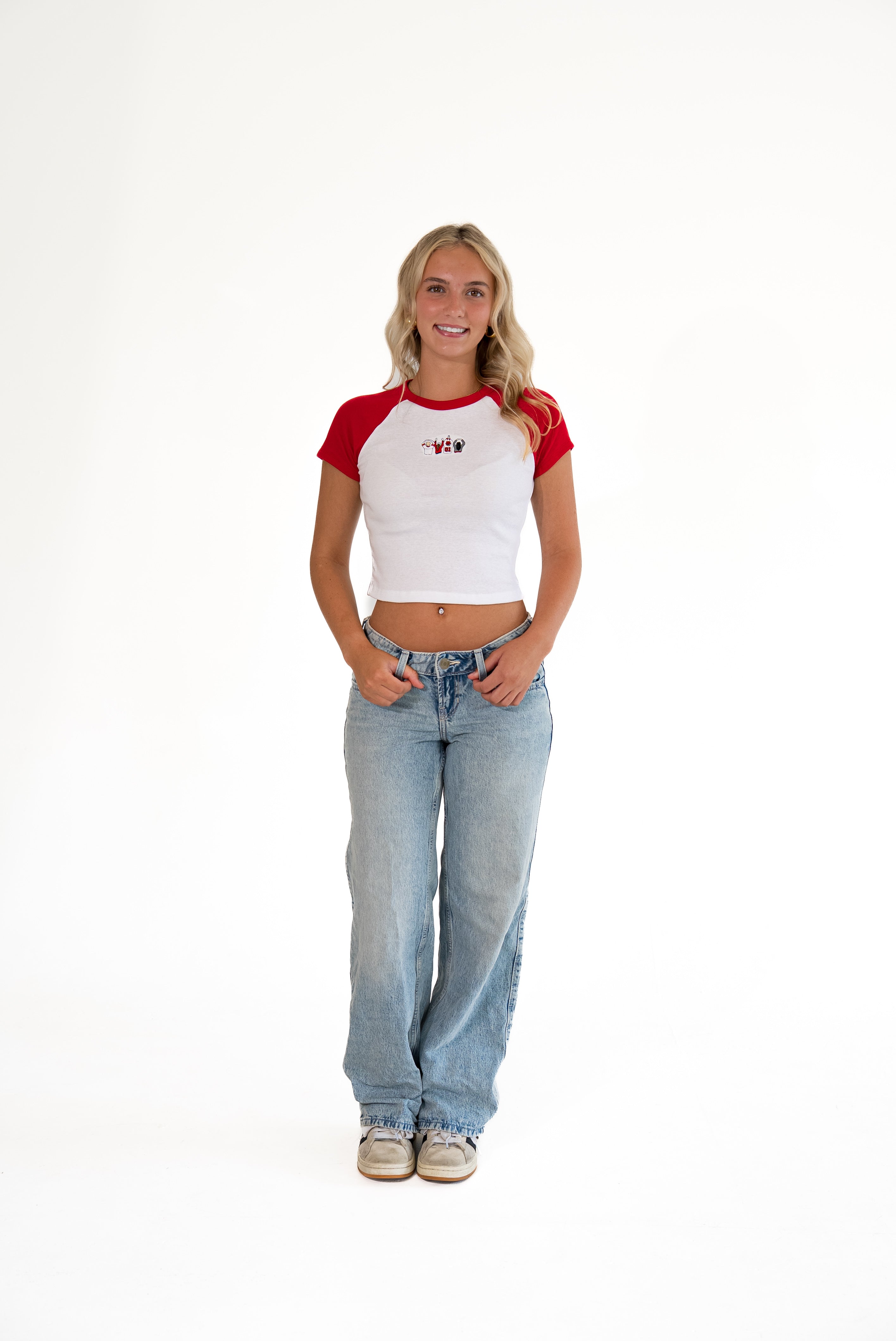 A young woman stands against a plain white studio background wearing a white and red raglan crop top with a small red graphic on the chest, light-wash blue jeans, and white sneakers.