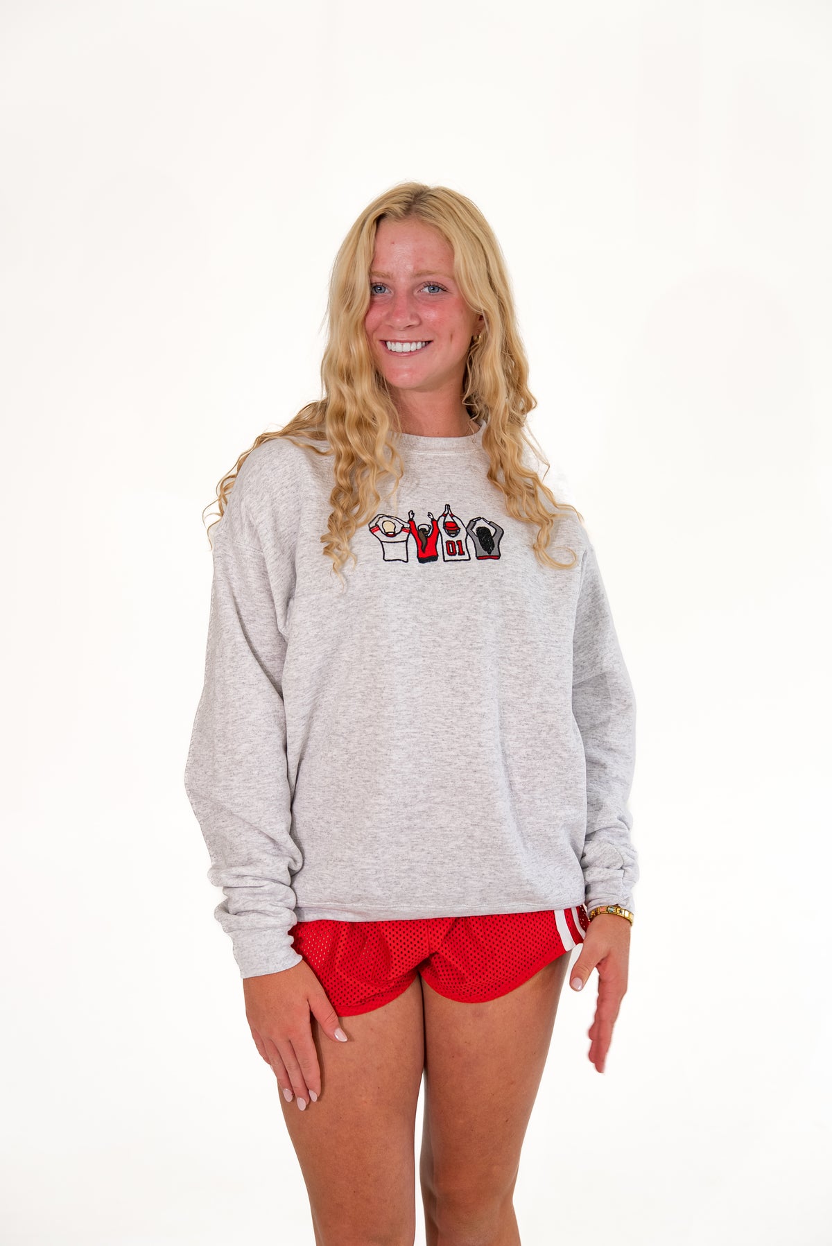 A smiling blonde woman wearing a light gray heather crewneck sweatshirt with a small graphic on the chest and red athletic shorts, standing against a plain white studio background.
