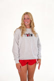 A smiling blonde woman wearing a light gray heather crewneck sweatshirt with a small graphic on the chest and red athletic shorts, standing against a plain white studio background.