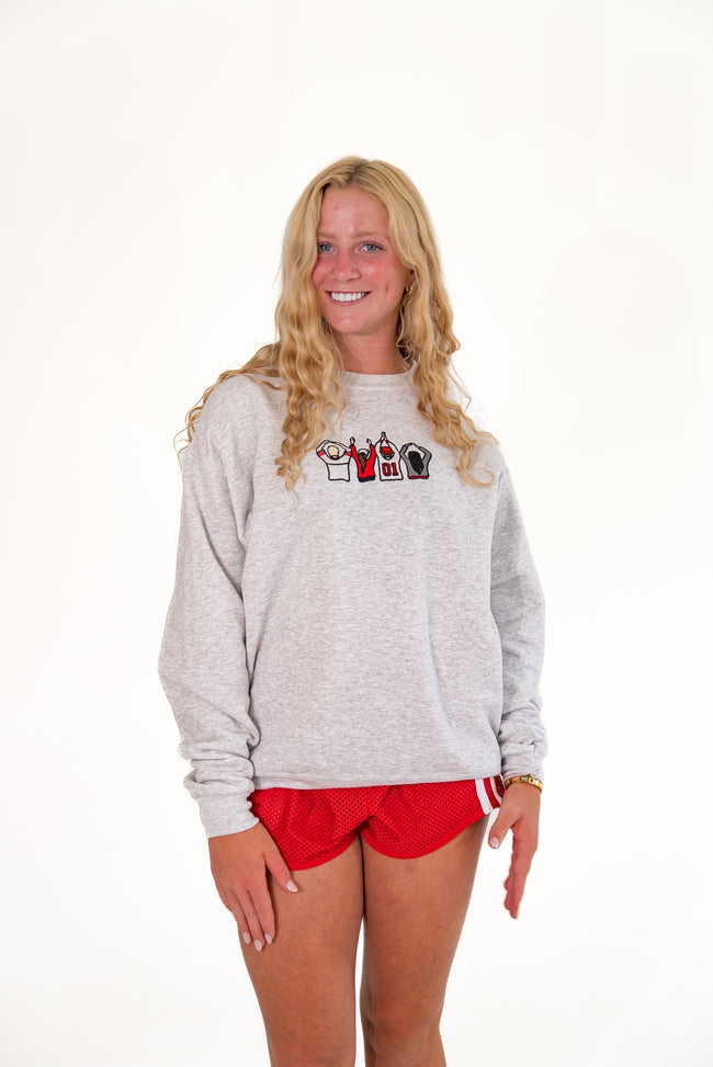 A smiling blonde woman wearing a light gray heather crewneck sweatshirt with a small graphic on the chest and red athletic shorts, standing against a plain white studio background.