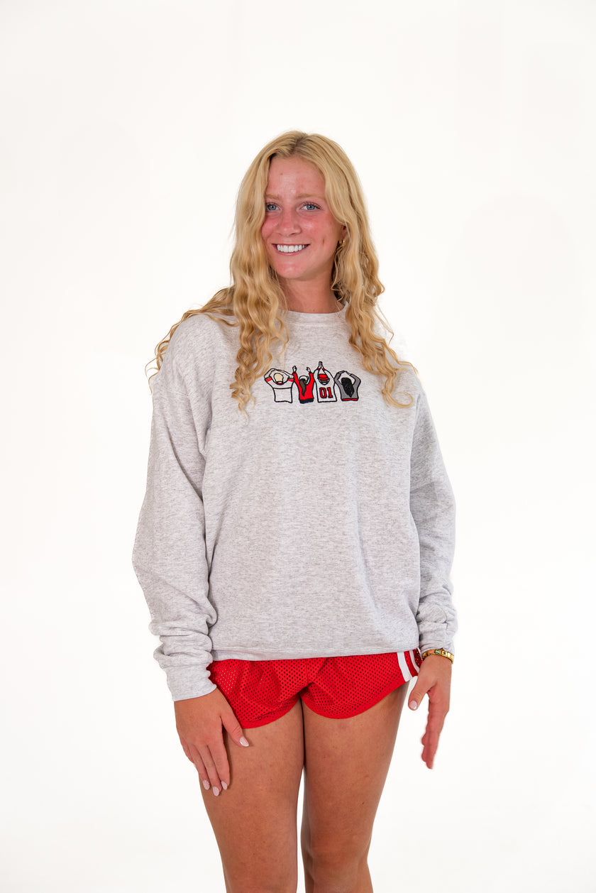 A smiling blonde woman wearing a light gray heather crewneck sweatshirt with a small graphic on the chest and red athletic shorts, standing against a plain white studio background.
