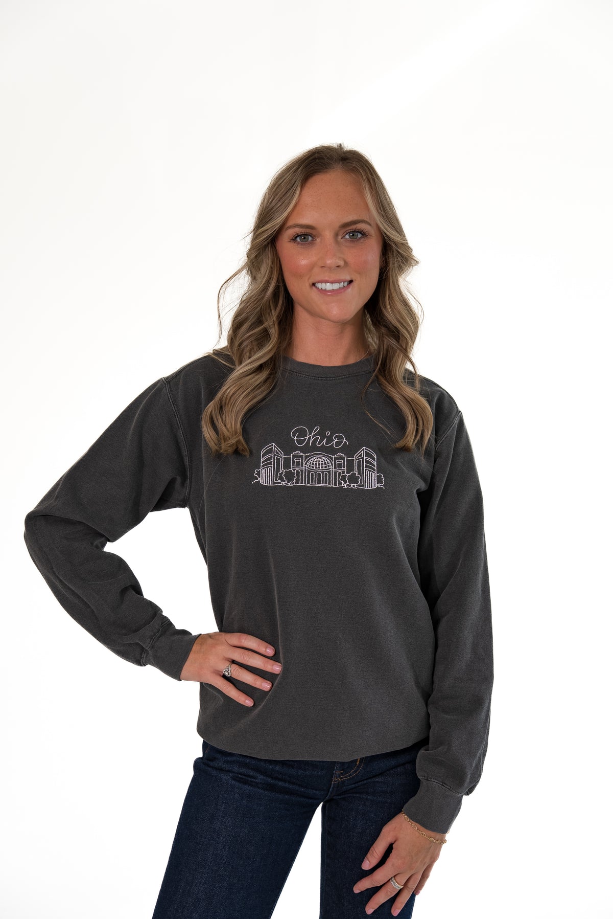 Woman standing against a white background with her hand on her hip, wearing a charcoal gray crewneck sweatshirt with an embroidered Ohio Stadium graphic, paired with dark wash jeans