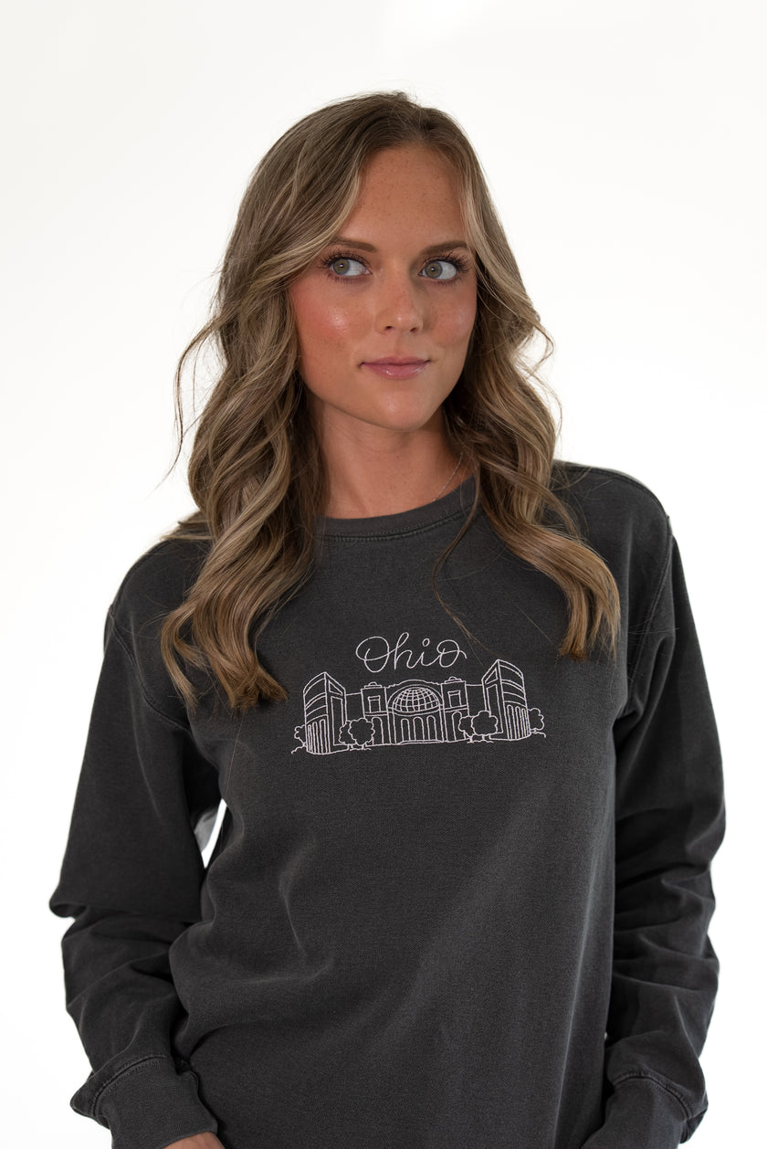 Close-up portrait of a woman looking away, wearing a charcoal gray crewneck sweatshirt featuring a fine white embroidered outline of the Ohio Stadium and script 