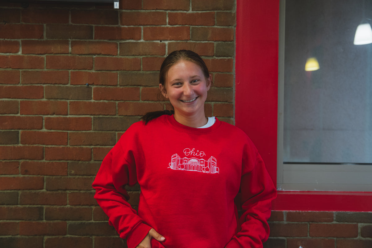 A smiling woman with her hair in a ponytail stands in front of a brick wall wearing a bright red sweatshirt with a white graphic of the Ohio state outline and the word 'Ohio' across the chest.