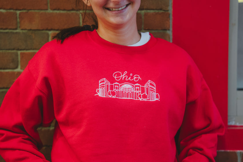 A person wearing a bright red crewneck sweatshirt with a white line art graphic of the Ohio state outline and the word 'Ohio' across the chest, standing in front of a brick wall and red door.