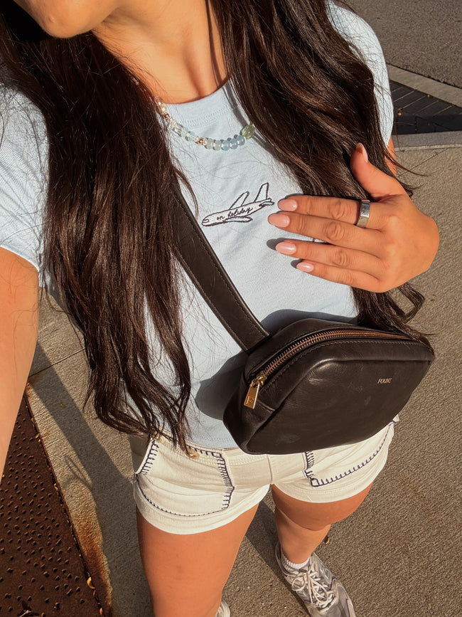 Woman wearing a light blue fitted baby tee with an embroidered airplane and “on holiday” lettering, styled with white shorts and a crossbody bag.