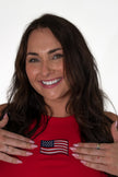 Close-up of a smiling woman gesturing toward a detailed embroidered American flag patch on the chest of a bright red ribbed tank top.