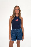 Smiling blonde woman standing against a white background, wearing a navy blue high-neck ribbed tank top with an embroidered American flag patch, paired with patterned blue denim shorts with a frayed hem.