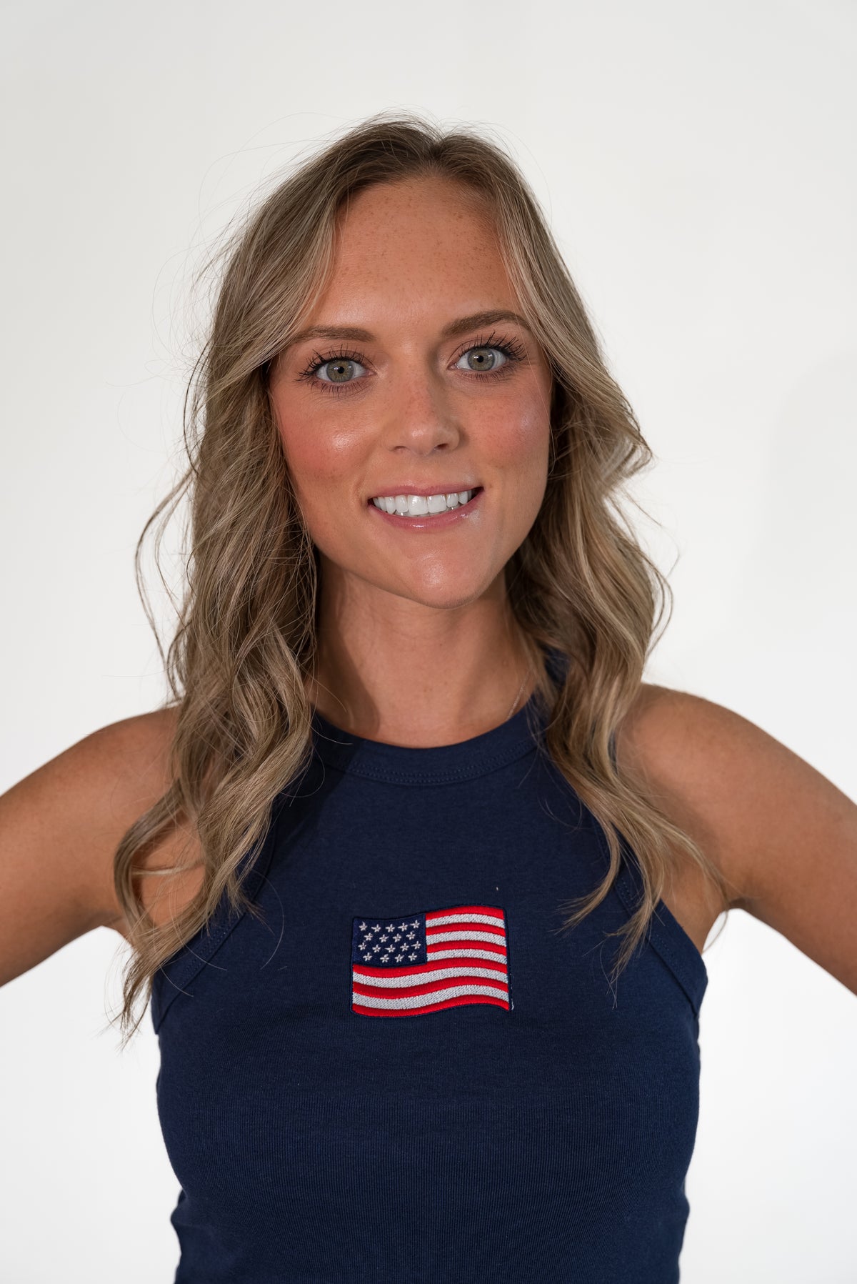 Close-up portrait of a smiling woman with blonde hair, wearing a navy blue high-neck ribbed tank top featuring an embroidered American flag patch.