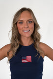 Close-up portrait of a smiling woman with blonde hair, wearing a navy blue high-neck ribbed tank top featuring an embroidered American flag patch.