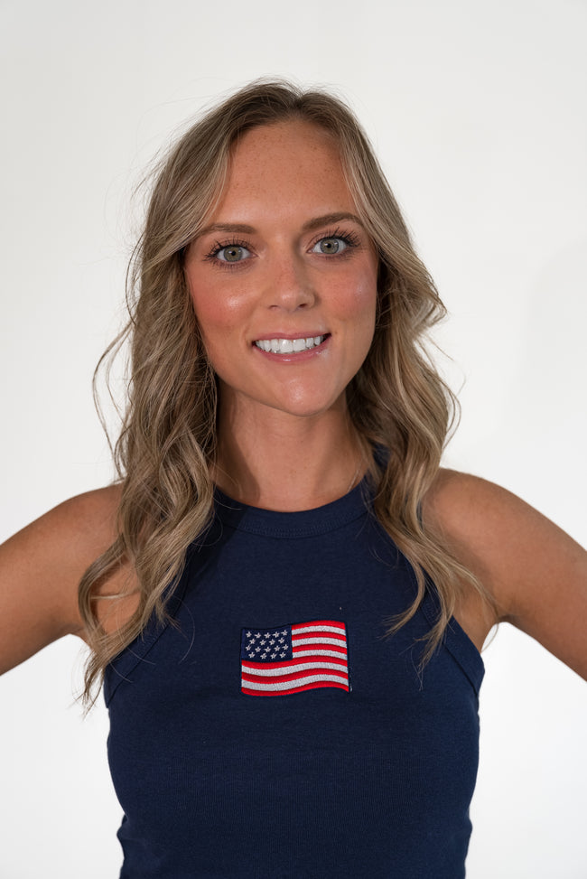 Close-up portrait of a smiling woman with blonde hair, wearing a navy blue high-neck ribbed tank top featuring an embroidered American flag patch.