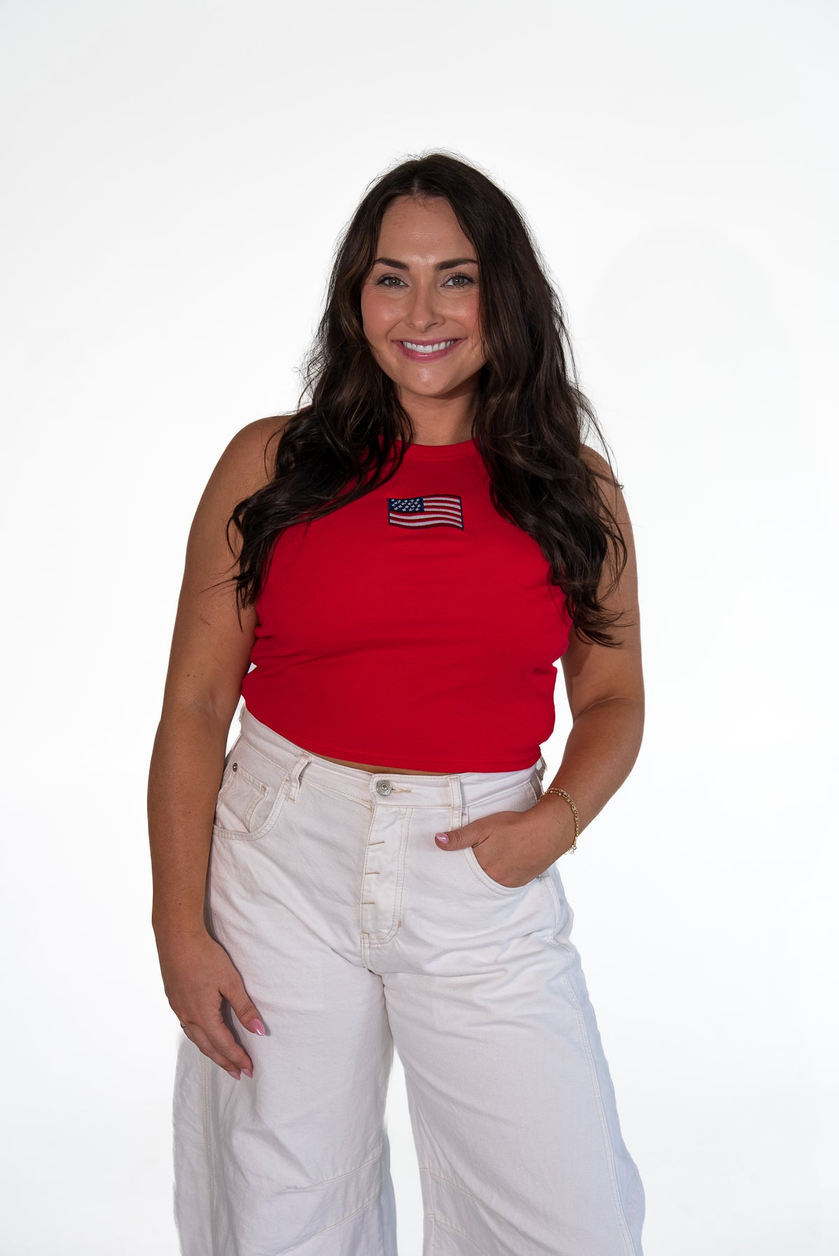 Smiling woman with long dark hair standing against a white background, wearing a bright red high-neck ribbed tank top with an American flag patch, paired with white wide-leg trousers.