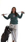 A woman stands on a white studio background holding a golf club over her shoulder next to a golf bag filled with clubs. She is wearing a dark green long-sleeve shirt with a pink and white graphic on the chest and white pants.