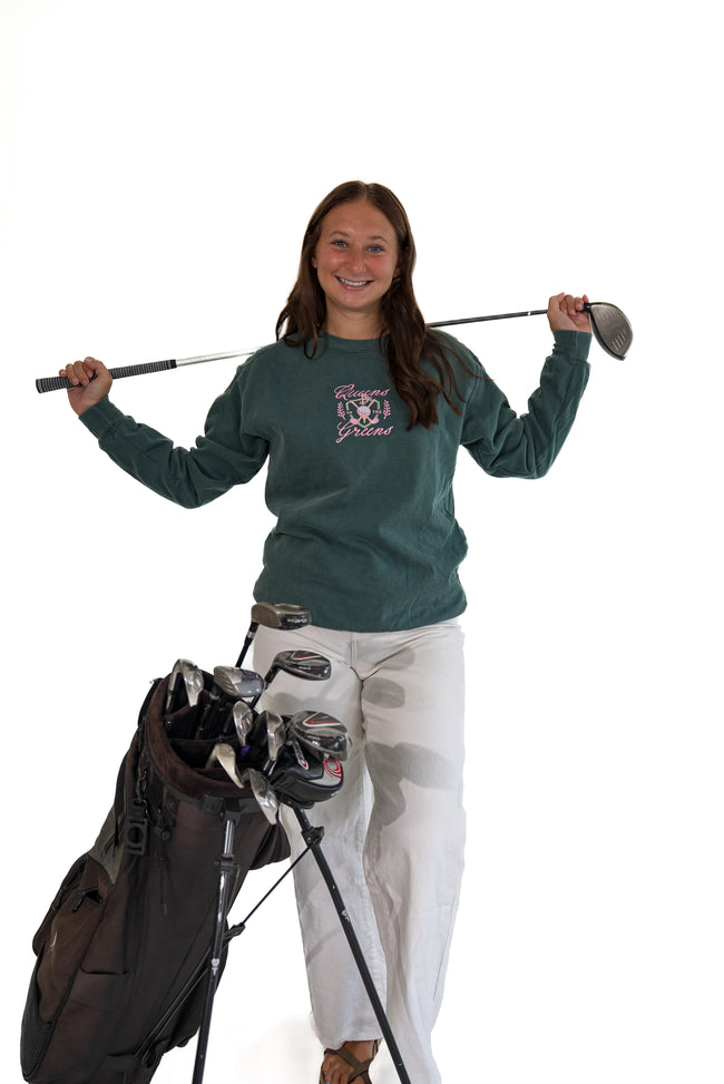 A woman stands on a white studio background holding a golf club over her shoulder next to a golf bag filled with clubs. She is wearing a dark green long-sleeve shirt with a pink and white graphic on the chest and white pants.