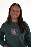 A smiling young woman with long brown hair wearing a dark teal-green crewneck sweatshirt with a pink and white 'Queens of the Greens' graphic on the front, set against a plain white background.