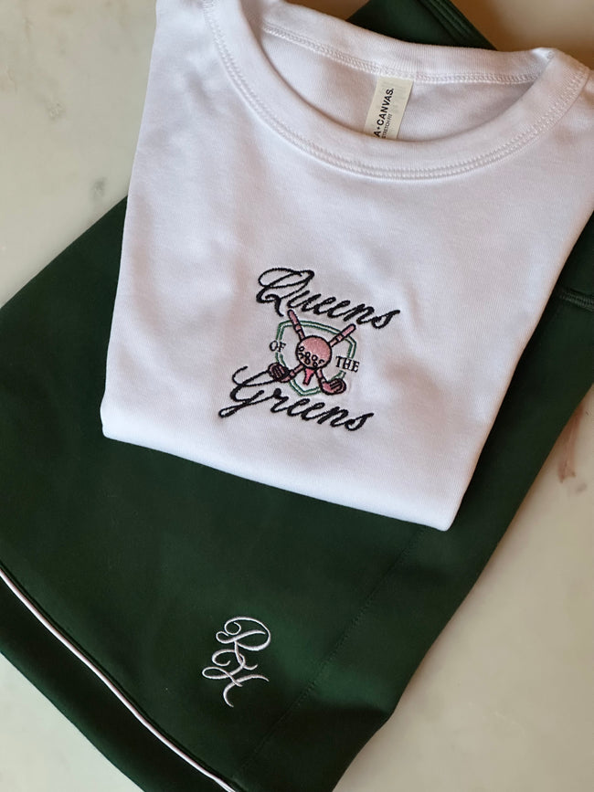 White fitted baby tee with embroidered “Queens of the Greens” lettering, golf clubs, and golf ball design.