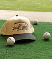 Golf trucker hat with embroidered “Girls Can Golf Too” and “golf club” lettering, displayed on turf with golf balls.