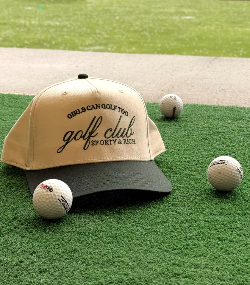 Golf trucker hat with embroidered “Girls Can Golf Too” and “golf club” lettering, displayed on turf with golf balls.