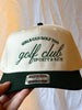 Golf trucker hat with embroidered “Girls Can Golf Too” and “golf club” lettering, displayed on turf with golf balls.